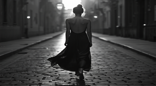 A woman walking down a cobblestone street in a flowing backless evening dress, shown from behind in classic black and white editorial style.
