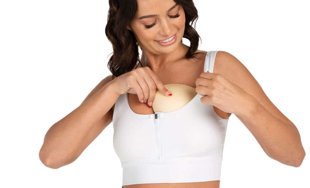 A dark hair female inserting the nude skin colour sports bra inserts whilst wearing  her white sports bra. The image is on a white background. 